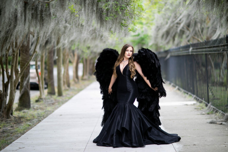 A woman in a black dress with black wings posing on a sidewalk.
