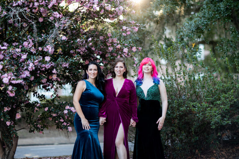Three women in evening gowns posing in front of a tree.